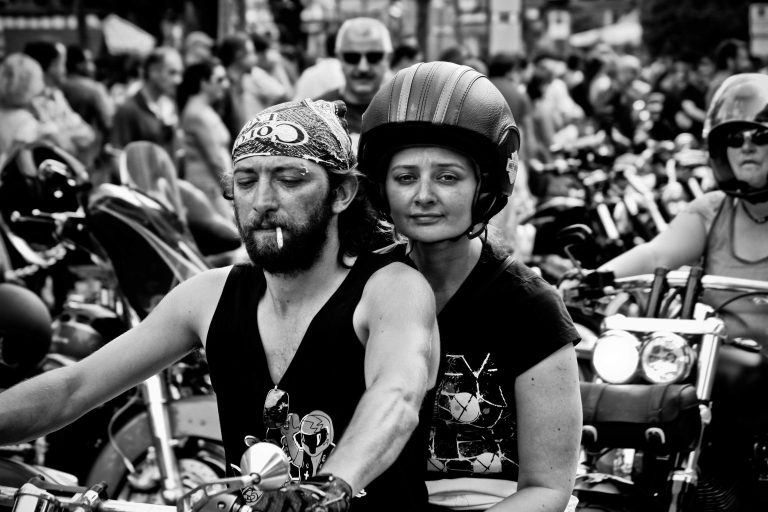 A couple ride on a bike in vests during a festival image is in black and white for Kutte Kulture LTD preserving outlaw motorcycle club legacy